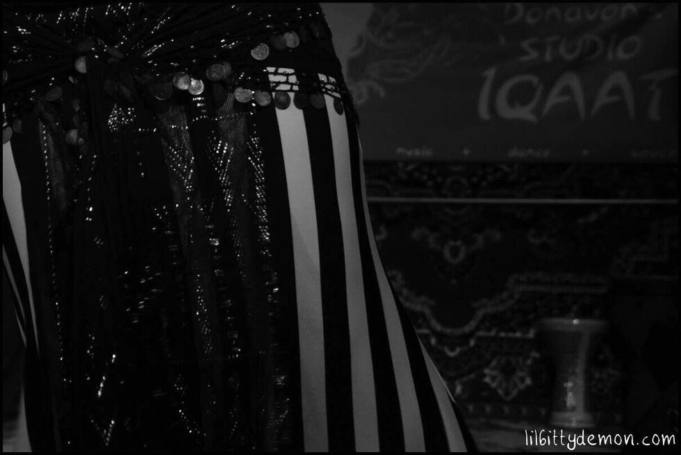 Black-and-white photo of a dancer’s coin belt and striped skirt in front of the Studio Iqaat banner, with a dumbek in the background